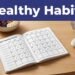 An open 30-day habit journal sits beside water, fruit, a meal bowl, sneakers, and a clock.