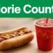 A hot dog and red soda cup sit on a wooden surface below a green headline.