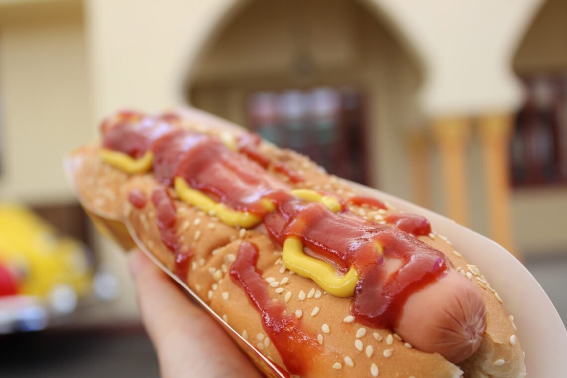 A hot dog on a white plate beside a red soda cup.