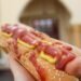 A hot dog on a white plate beside a red soda cup.